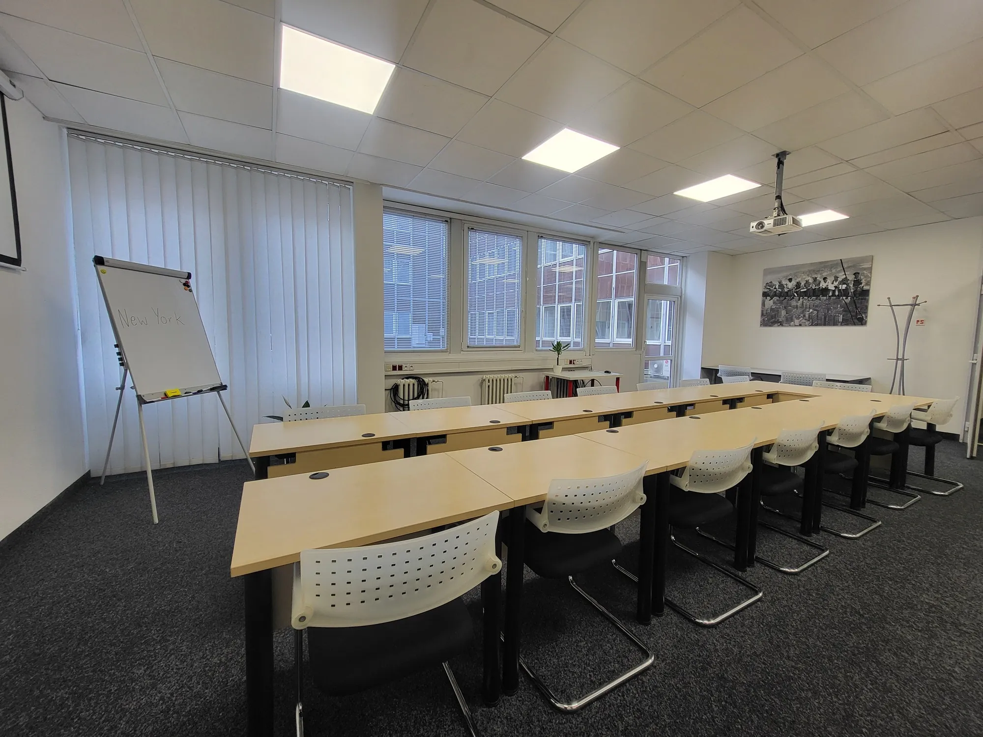 Modern conference room with U-shaped wooden tables, white chairs, a flip chart labeled 'New York', and windows with blinds.